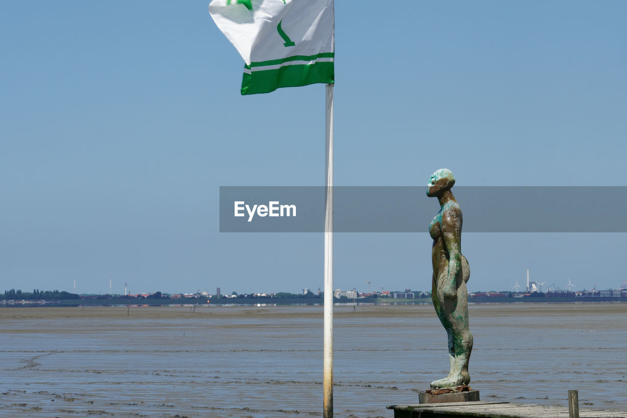 water, sky, nature, flag, sculpture, clear sky, day, patriotism, statue, sea, wind, beach, human representation, representation, land, environment, full length, outdoors, blue, sunny, architecture, copy space, no people, travel