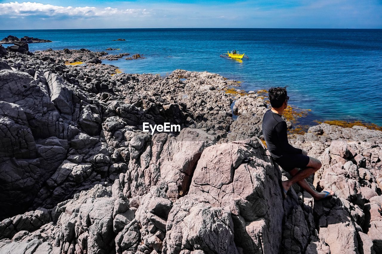 Rear view of man sitting by sea on rock during sunny day