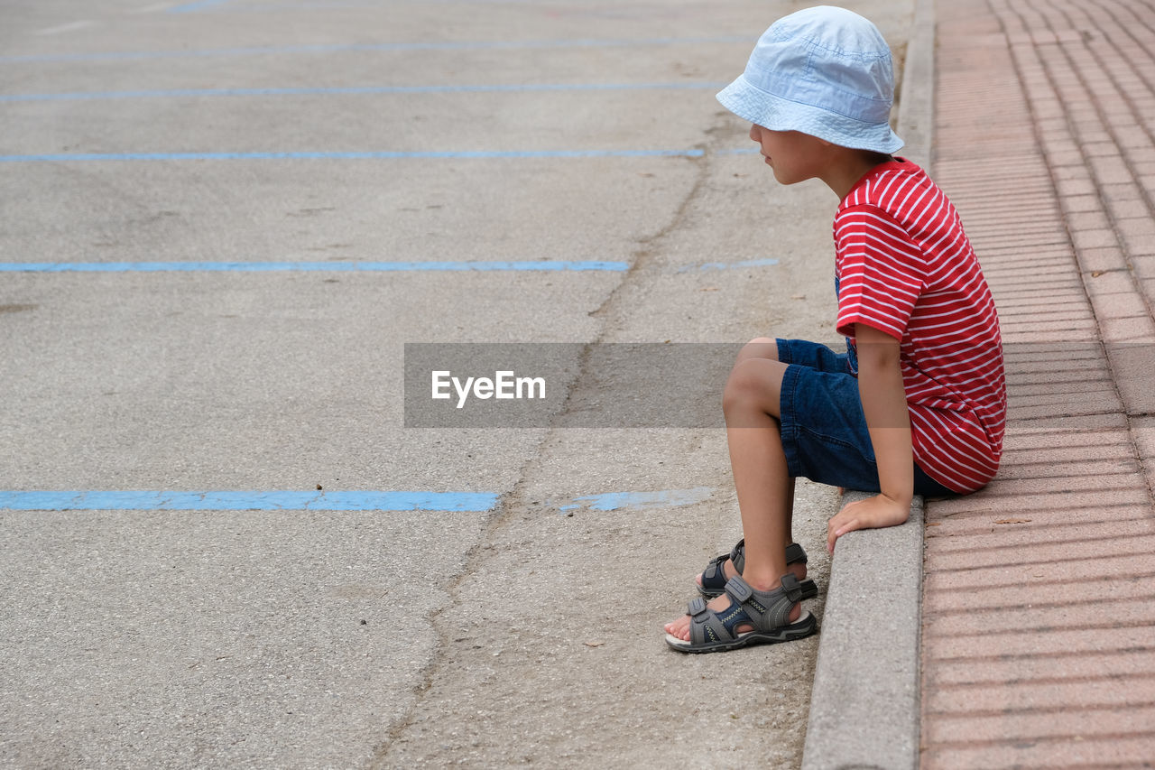 Side view of boy wearing hat sitting on footpath