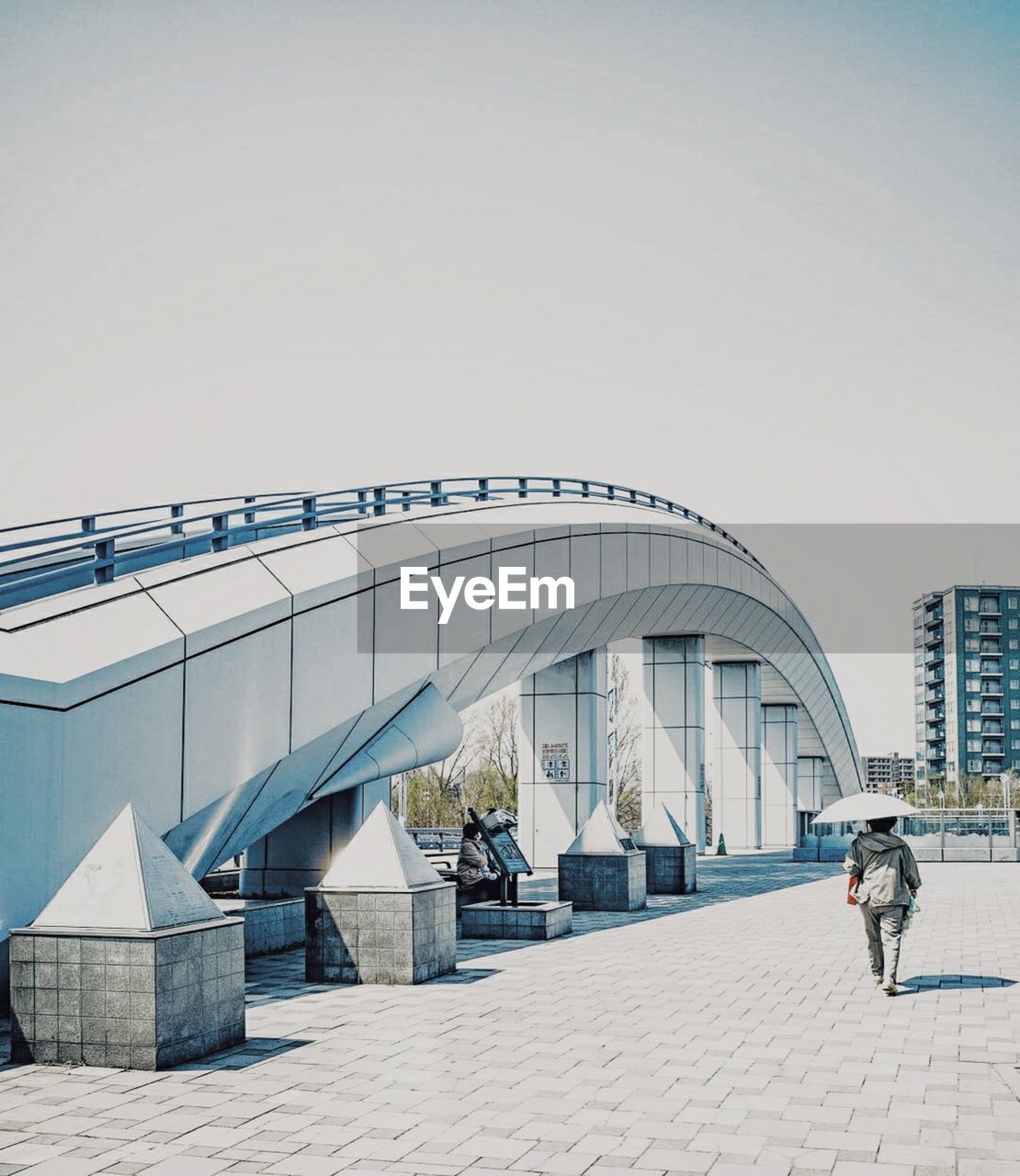 REAR VIEW OF PEOPLE WALKING ON BRIDGE AGAINST SKY IN CITY