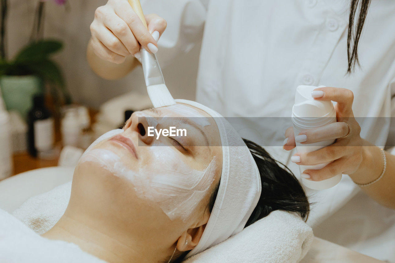 A girl cosmetologist applies a cream mask to a woman s face with a brush.