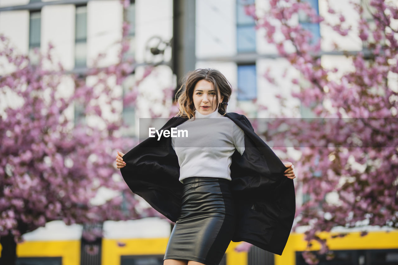 Portrait of beautiful woman holding winter coat against cherry trees in city