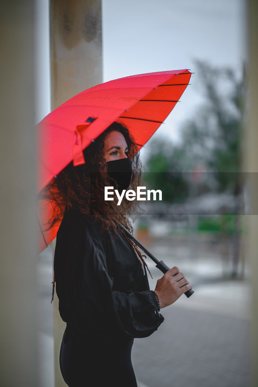 Woman with a mask and a red umbrella seen between two columns