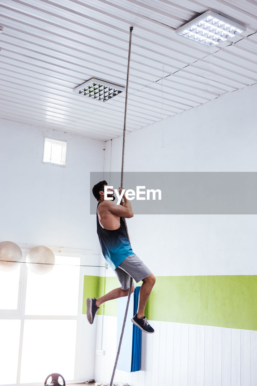 From below side view of young athletic male in sportswear performing rope climb exercise during fitness training in modern gym