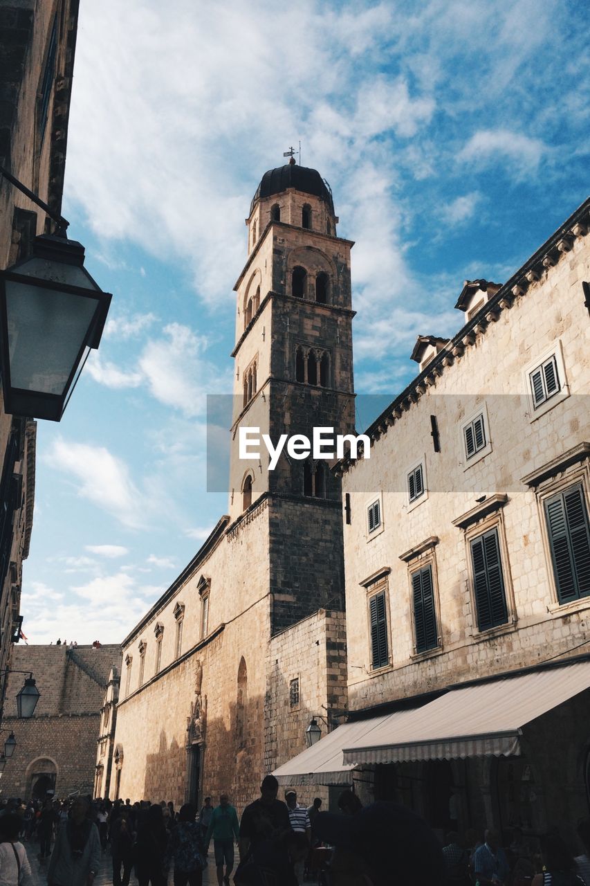 Low angle view of people walking on street in front of steeple against sky