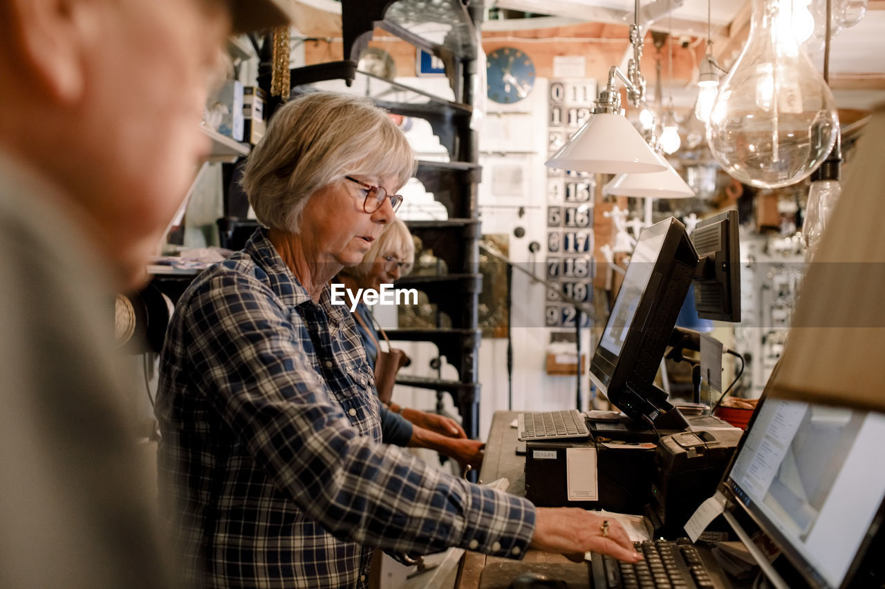 Confident senior female owner using computer while standing amidst coworkers at checkout in illuminated hardware store