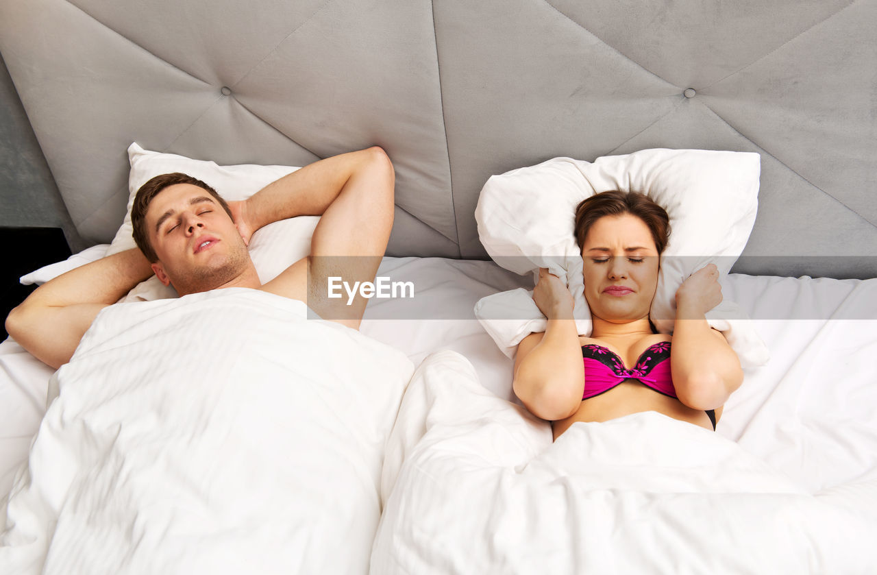 Young man sleeping while woman covering ears with pillow on bed at home