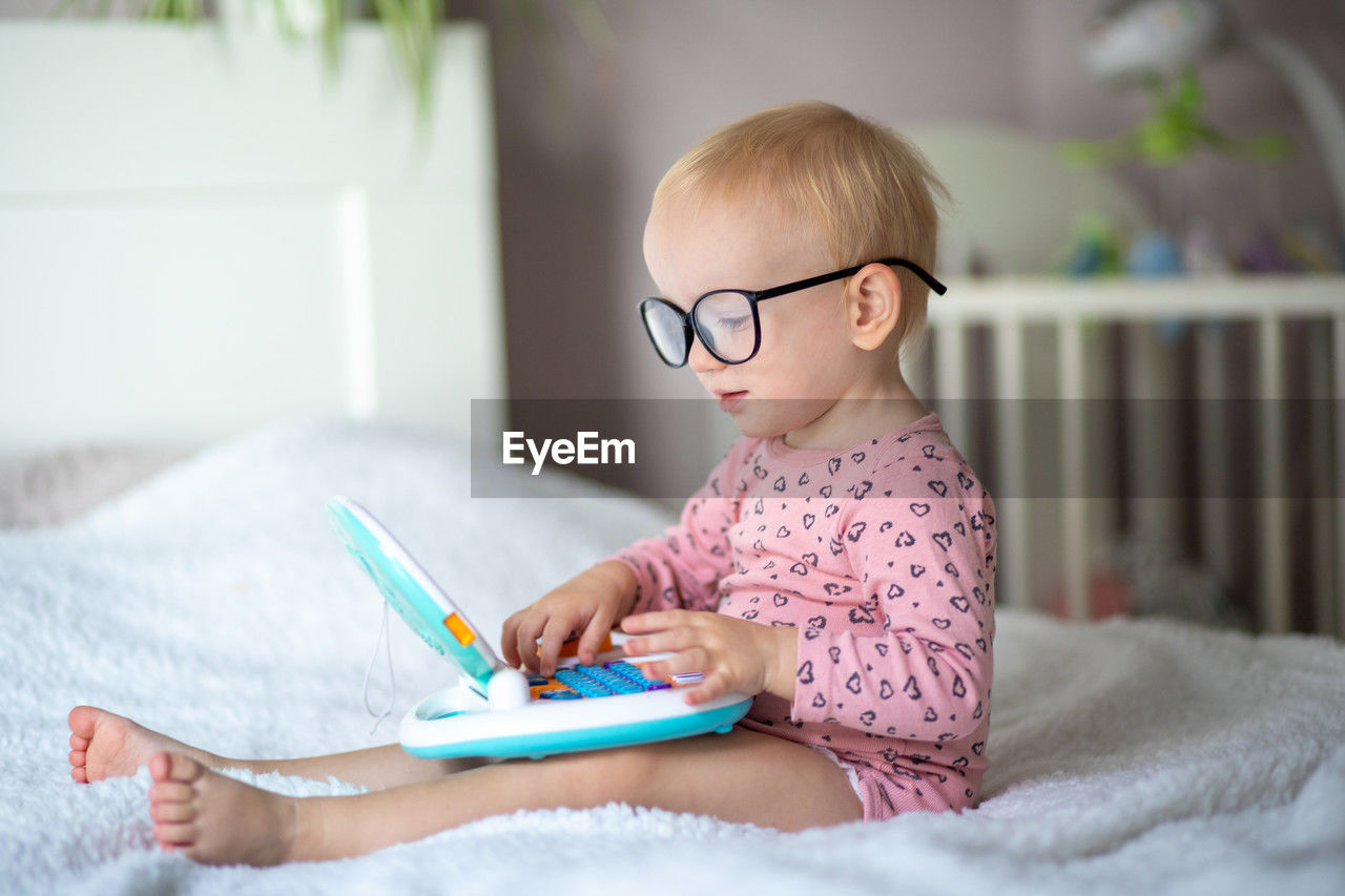 child, childhood, one person, indoors, toddler, domestic room, furniture, eyeglasses, sitting, baby, glasses, publication, innocence, bed, learning, domestic life, book, cute, lifestyles, person, home interior, casual clothing, bedroom, education, blond hair, women, activity, female, looking, clothing, relaxation, concentration, communication, smiling, baby clothing, holding, pink, happiness, reading, emotion, portrait, day, digital tablet