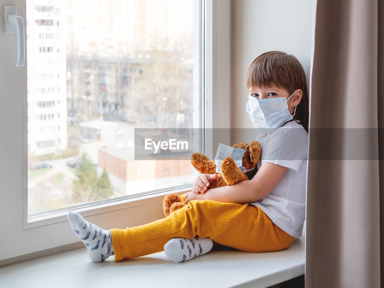 Toddler boy with teddy bear in medical masks looks through window.quarantine coronavirus covid-19.