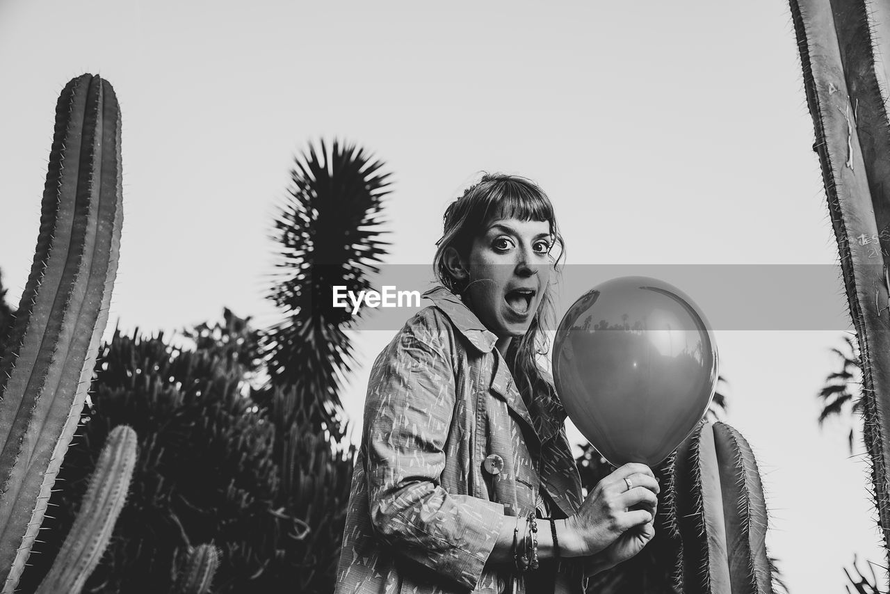 Low angle portrait of woman holding balloon while standing amidst cacti against clear sky