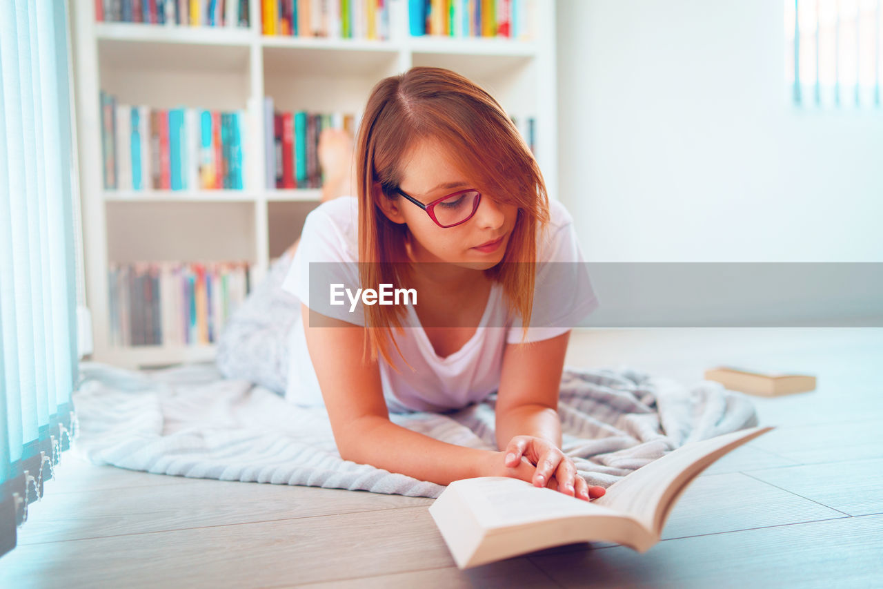 Beautiful woman wearing eyeglasses reading book lying on floor at home