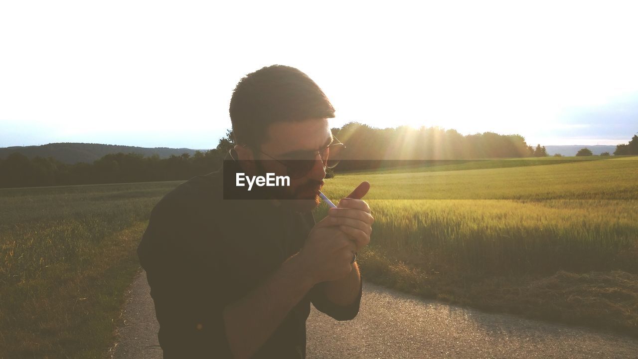 Man igniting cigarette while standing on grass against sky