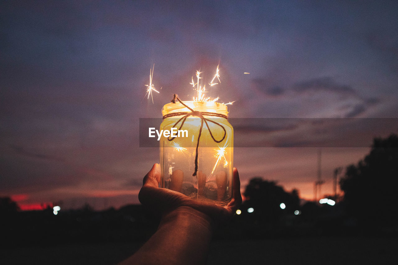 Cropped hand holding burning sparklers in container against sky at night