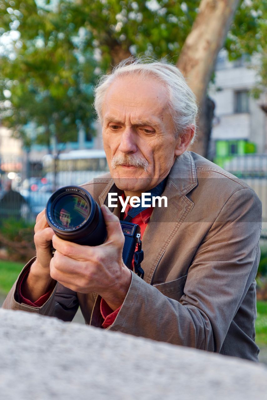 PORTRAIT OF MATURE MAN HOLDING CAMERA WHILE STANDING OUTDOORS