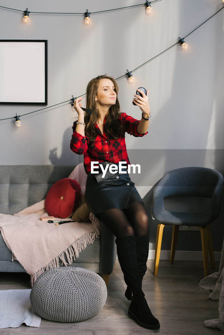 Young caucasian woman in a red plaid shirt and skirt does her makeup, paints blush with a brush 