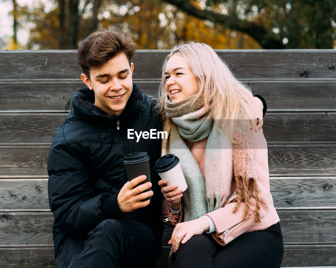 Cheerful young couple holding disposable coffee cups while sitting on bench at park