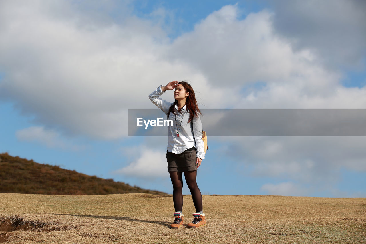 Full length of woman shielding eyes while standing on field against sky
