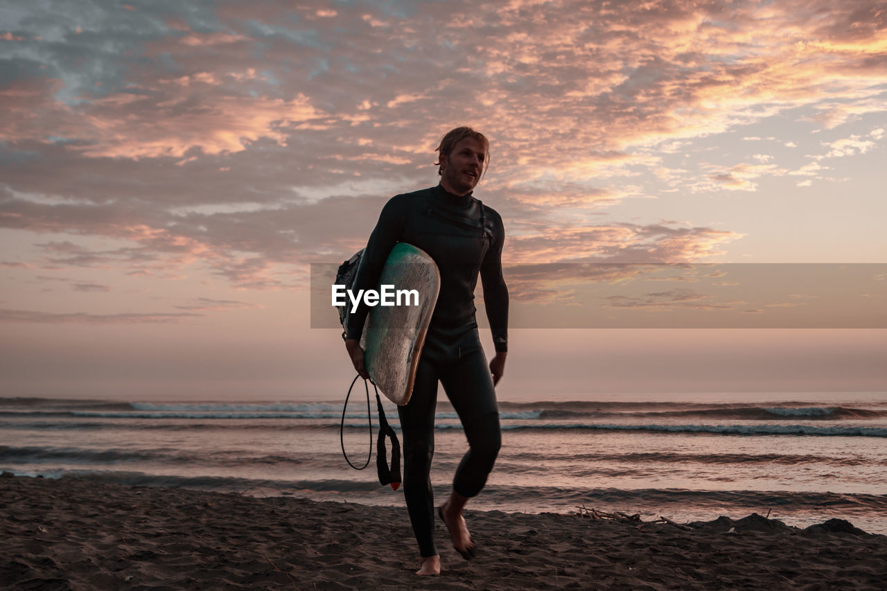Full length of man carrying surfboard at beach against sky during sunset