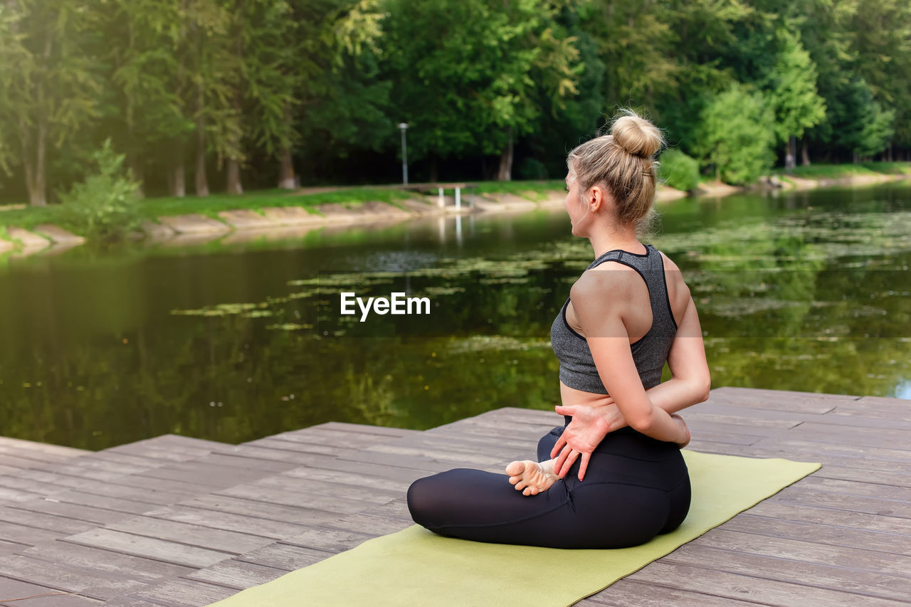 A woman sitting on a wooden platform by a pond in the park in summer and performing yoga