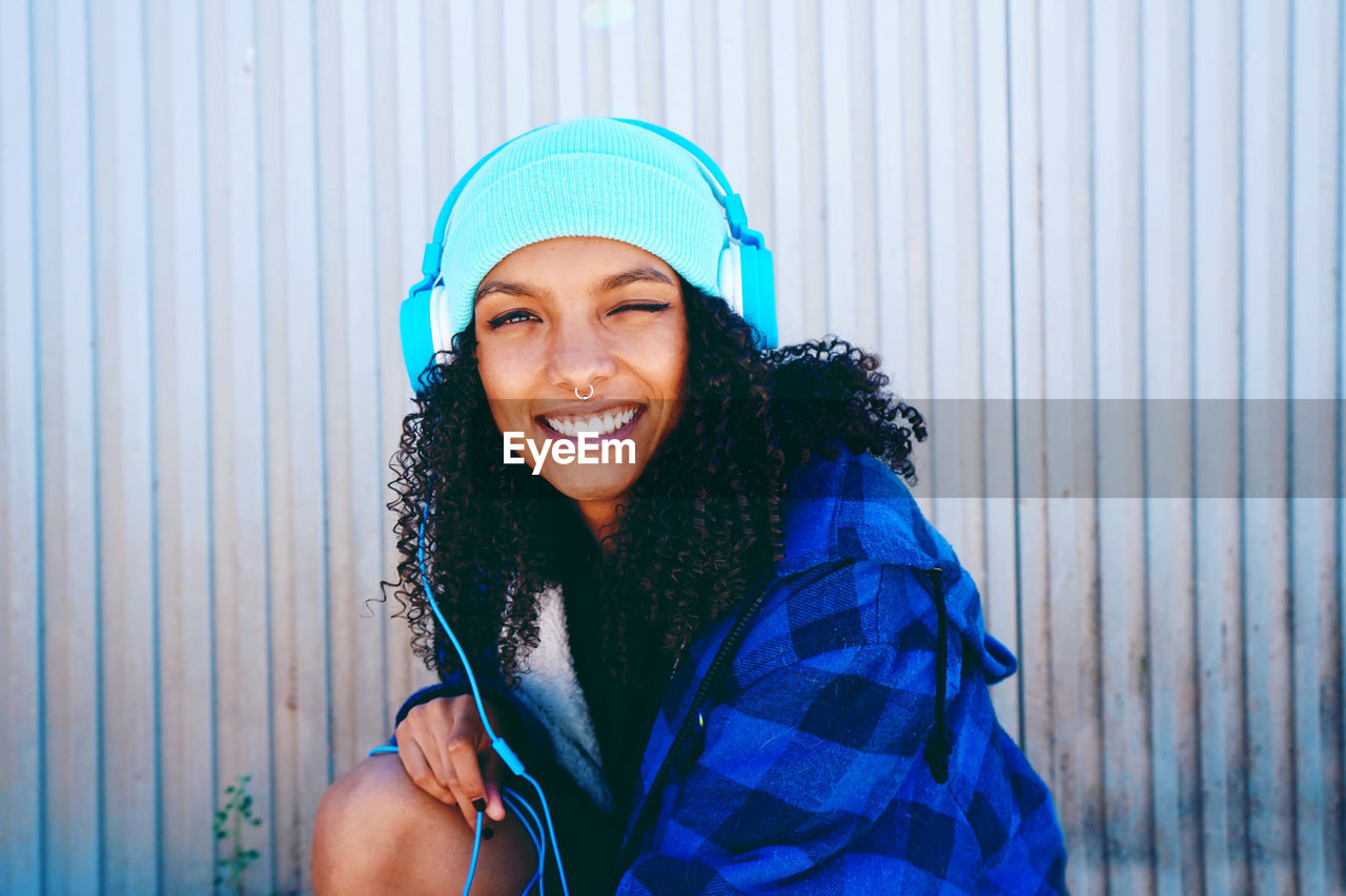 Portrait of smiling woman listening music while winking against wall