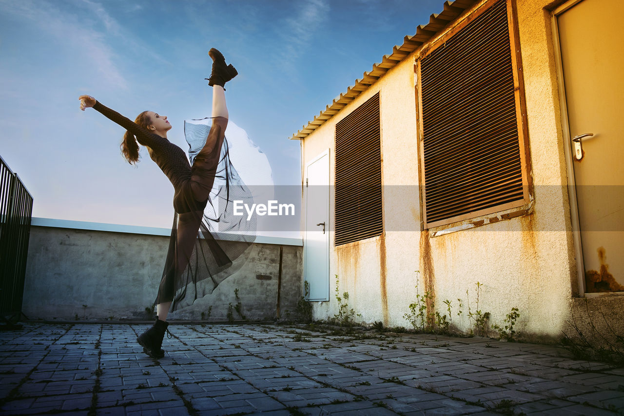 A ballerina in a black skirt  does vertical splits on the roof of a building against the sky