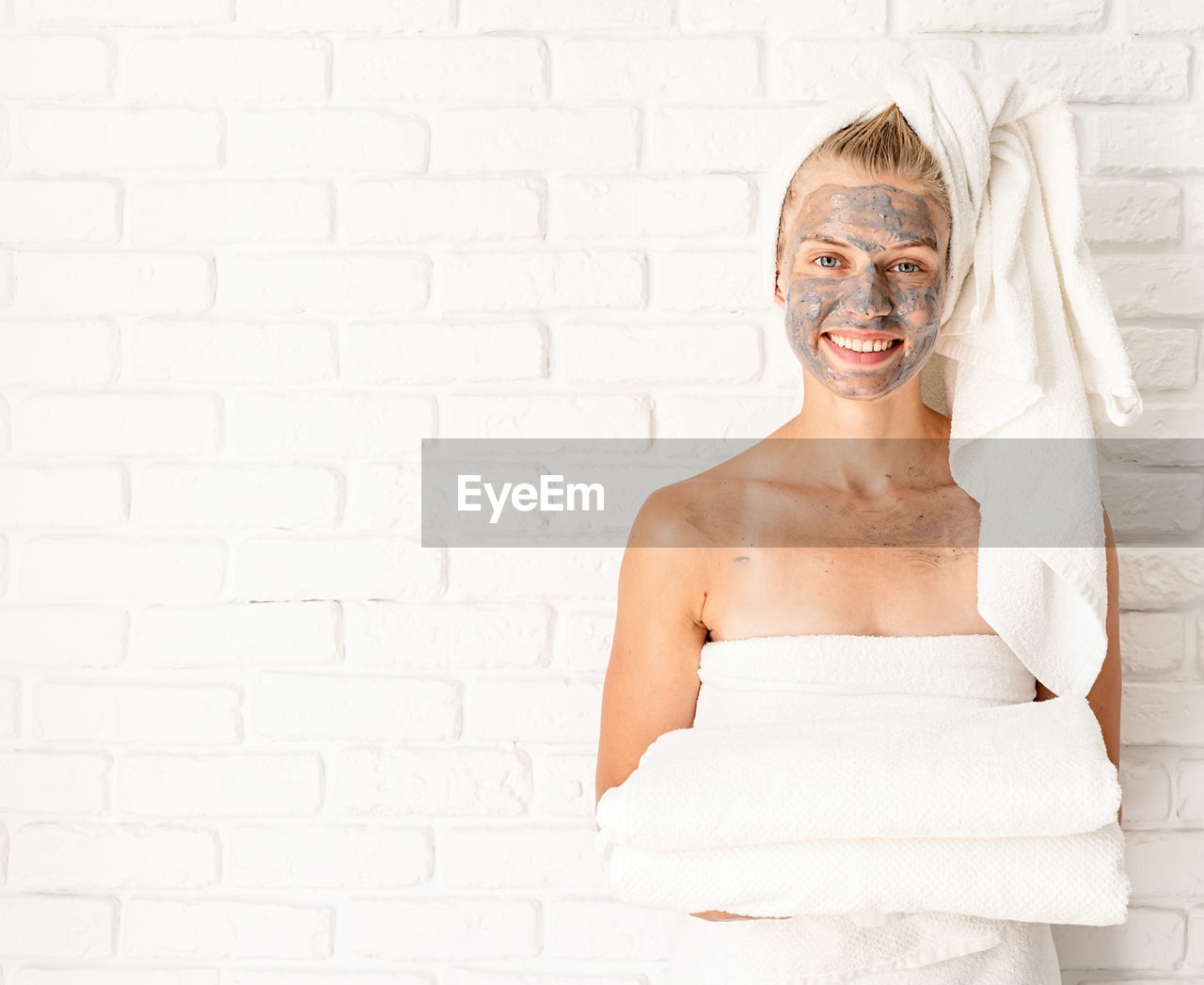 Spa facial mask. young smiling woman holding white bath towels with a clay facial mask on her face