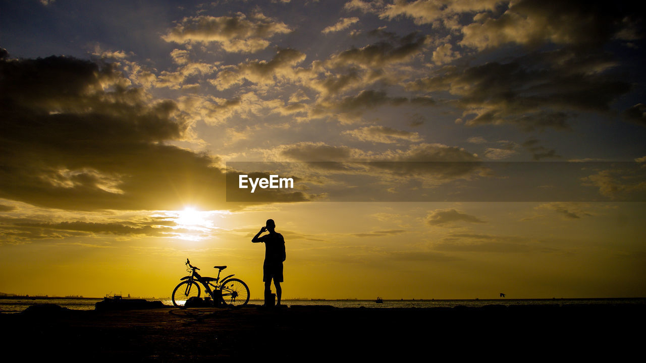 Silhouette man by bicycle against cloudy sky during sunset