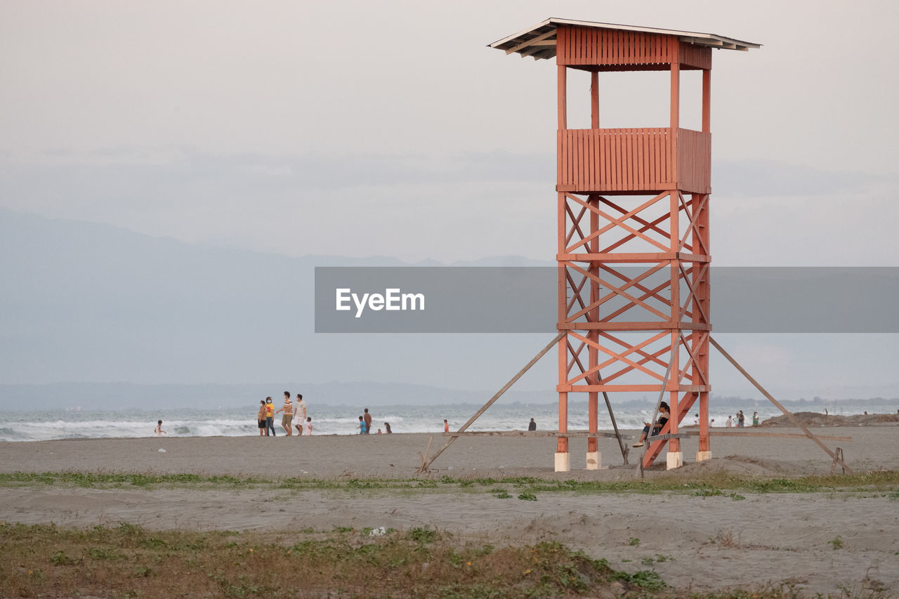 tower, beach, water, sea, land, sky, lifeguard hut, architecture, built structure, nature, man made structure, coast, lighthouse, lifeguard, observation tower, sand, group of people, scenics - nature, hut, day, beauty in nature, lookout tower, travel destinations, protection, observation point, men, outdoors, building exterior, security, environment, coastline, travel, cloud, tourism, trip, vacation, ocean, holiday, horizon, tranquility, horizon over water, non-urban scene, adult