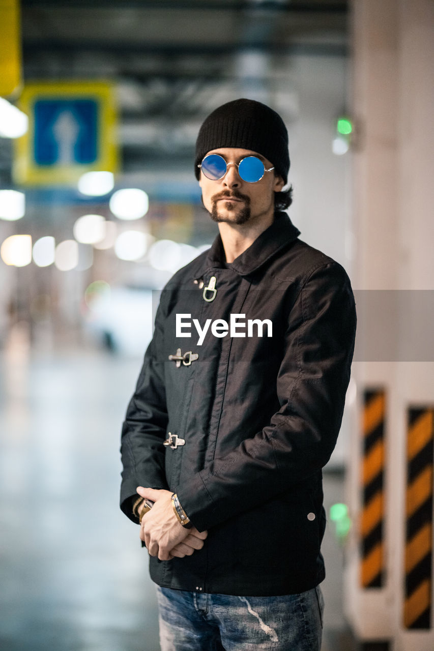 PORTRAIT OF YOUNG MAN WEARING SUNGLASSES STANDING AGAINST BLURRED BACKGROUND