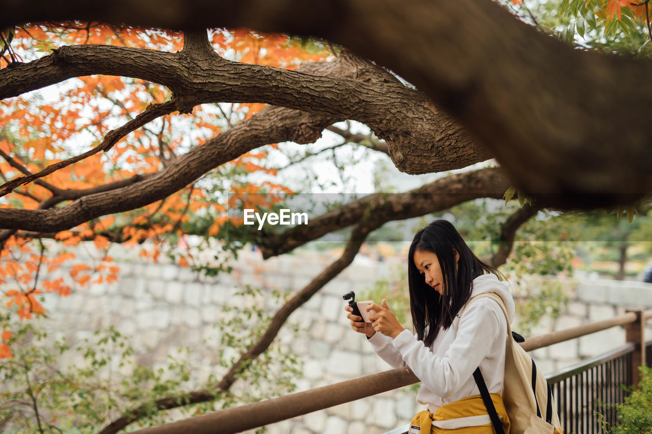 Portrait of young woman taking photo using mobile phone under a tree at the riverside 