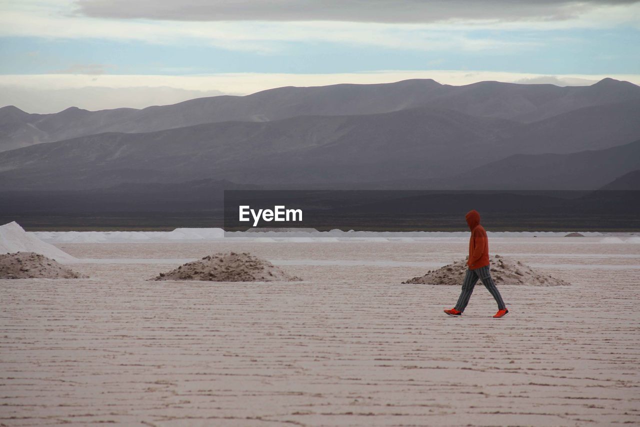 FULL LENGTH REAR VIEW OF MAN WALKING ON SHORE AGAINST MOUNTAINS