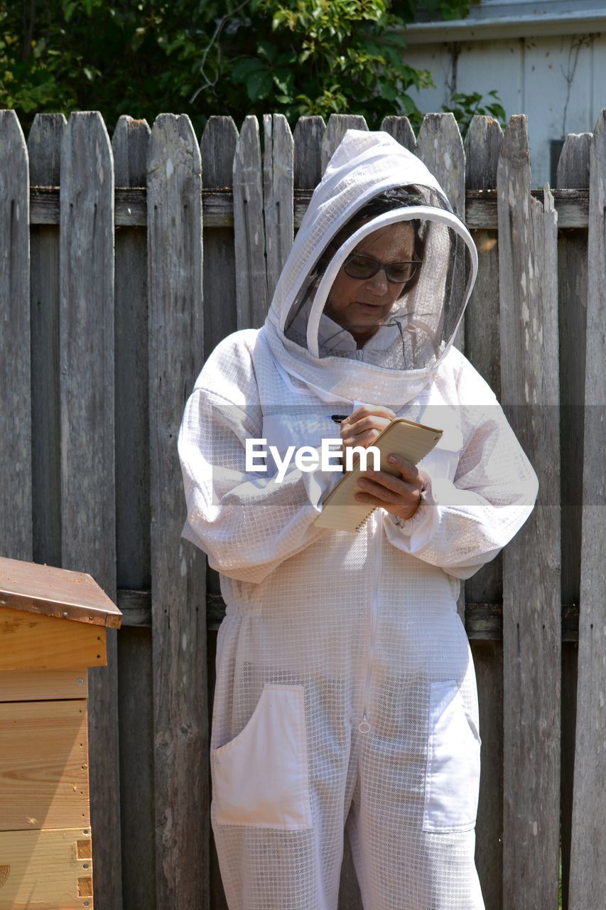 Woman writing in paper while standing by wooden fence