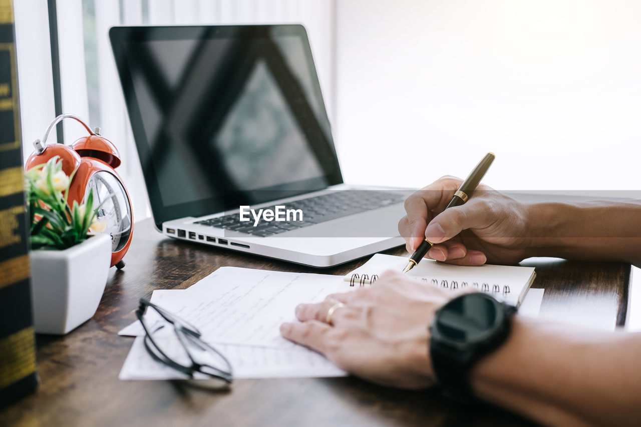 Cropped hands of man writing in book by laptop on table