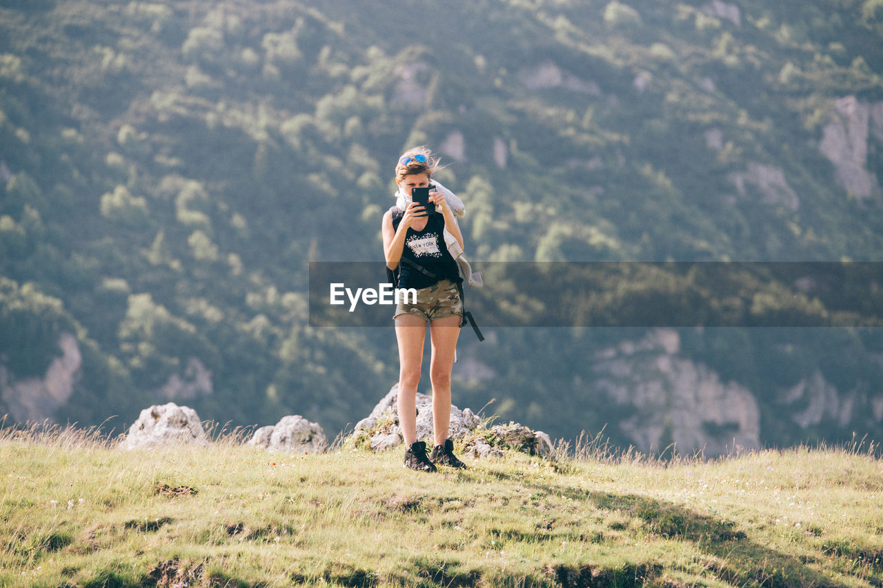 Full length of female hiker photographing while standing on field against mountain