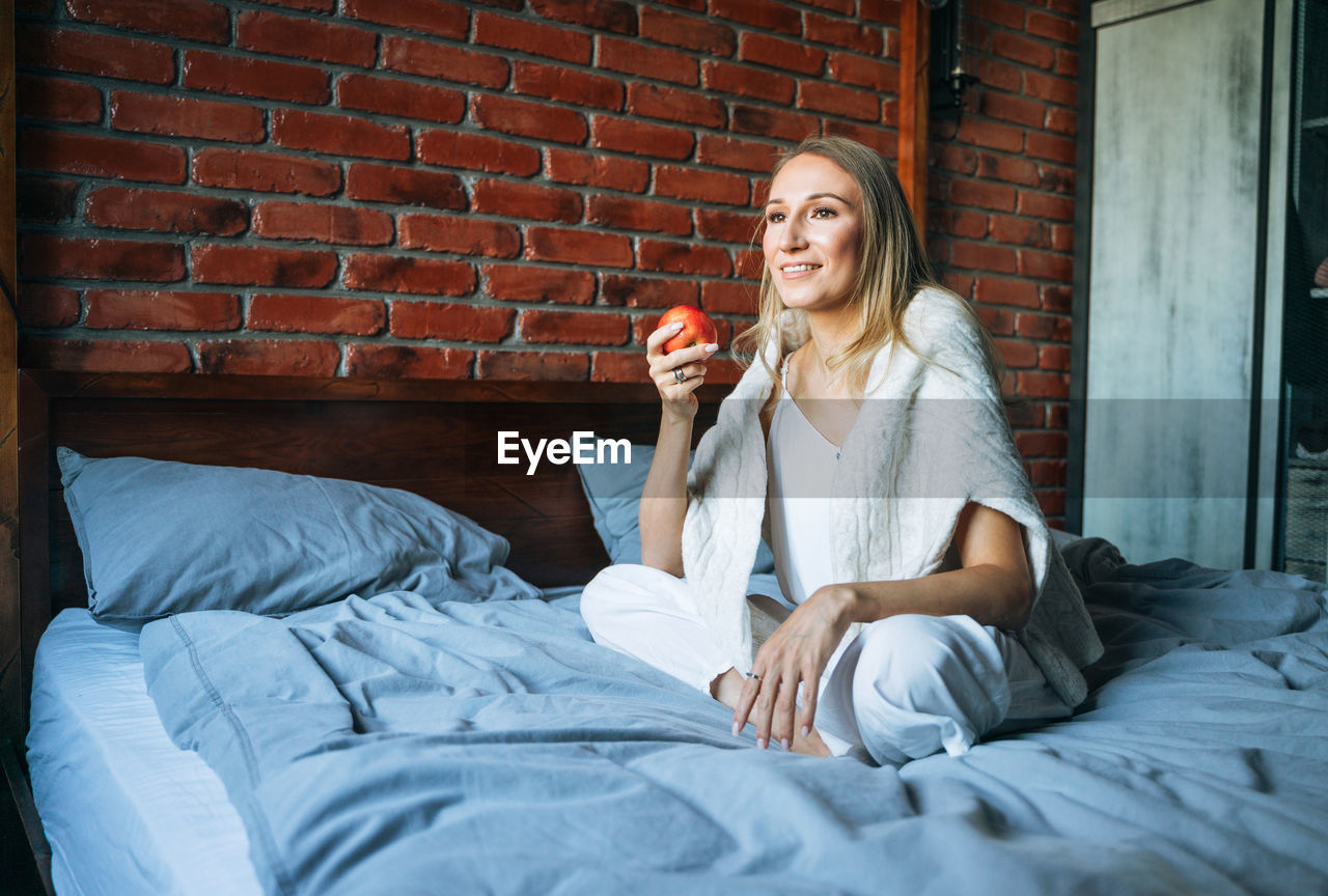 Young adult woman with blonde long hair in casual with apple fruit sitting on bed at home
