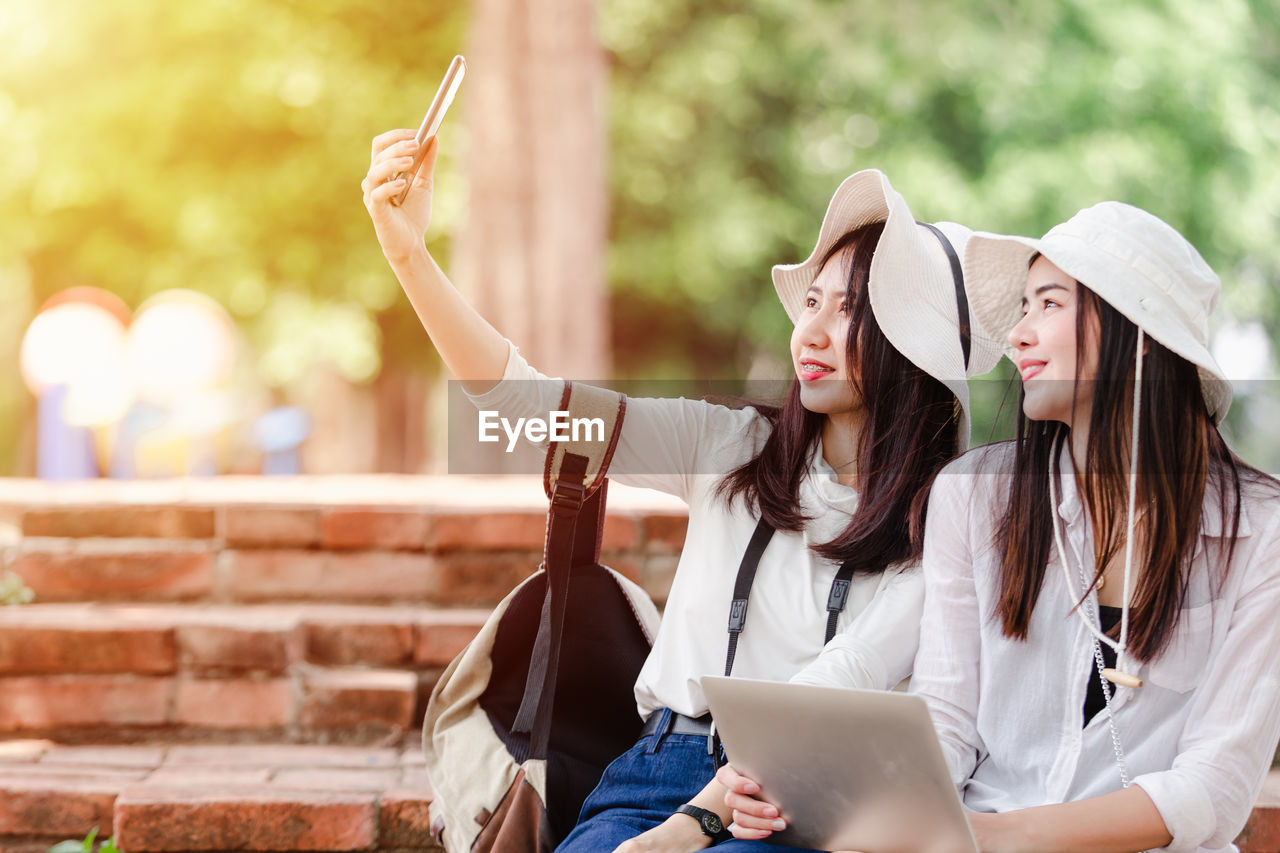 Woman talking selfie on smart phone with friend holding laptop