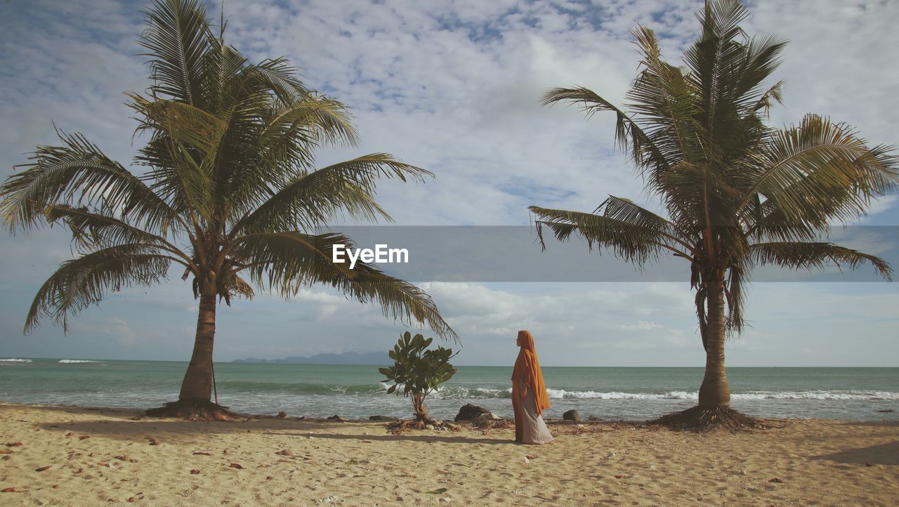 Palm trees on beach against sky with a muslim woman wearing hijab in between