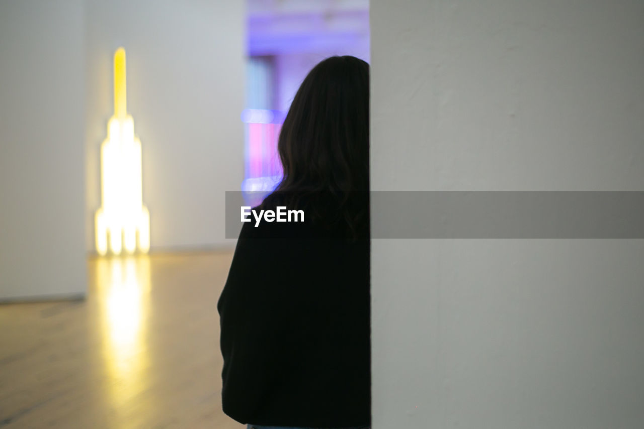 REAR VIEW OF WOMAN STANDING AGAINST WALL IN BUILDING