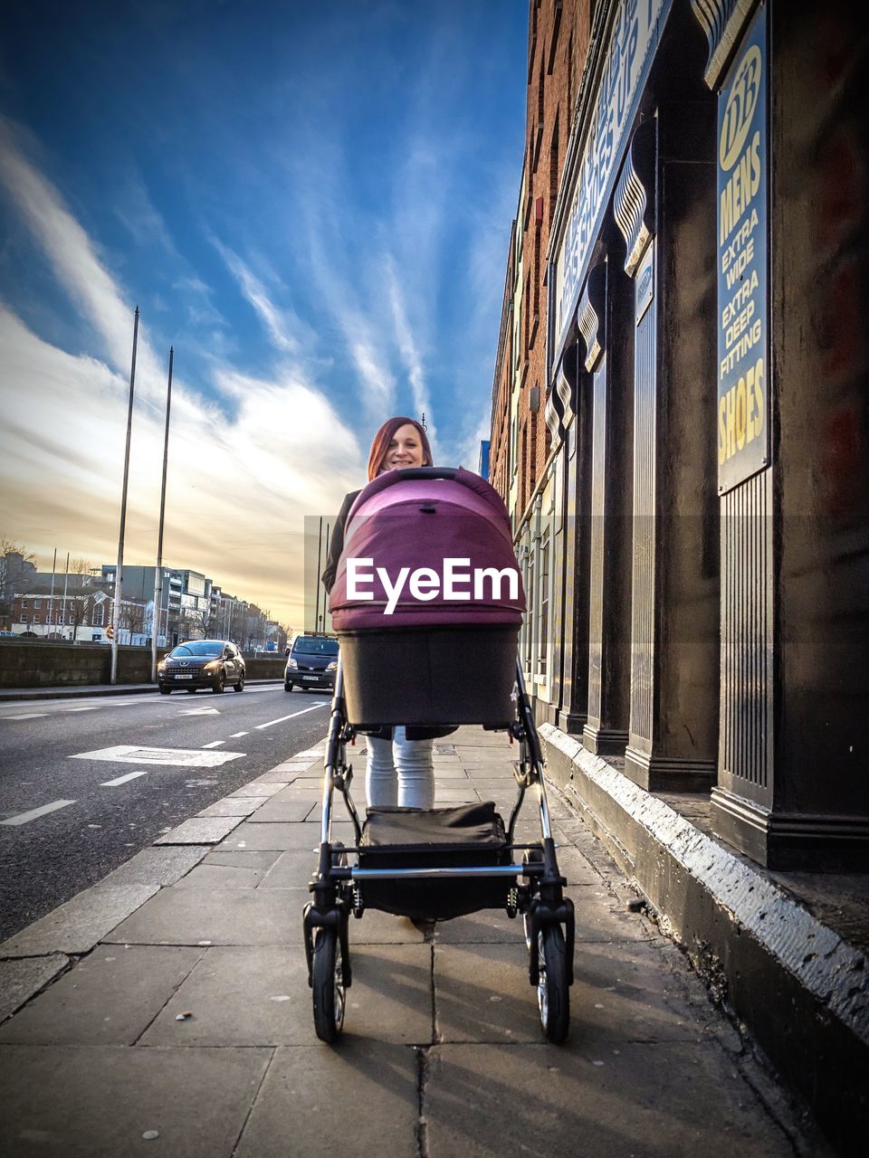 Smiling woman with baby carriage walking on sidewalk in city against sky