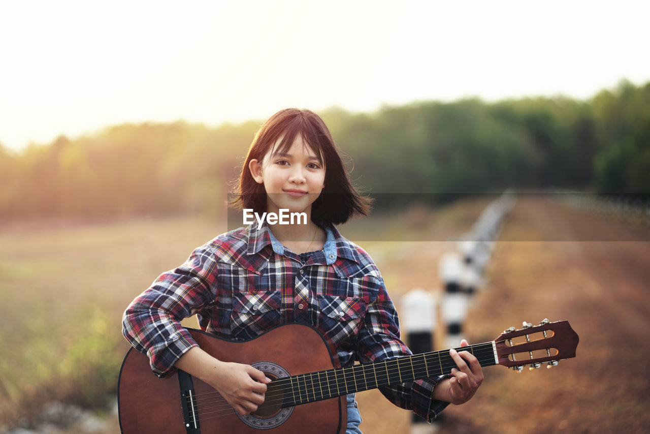 PORTRAIT OF YOUNG WOMAN PLAYING GUITAR AGAINST BLURRED BACKGROUND