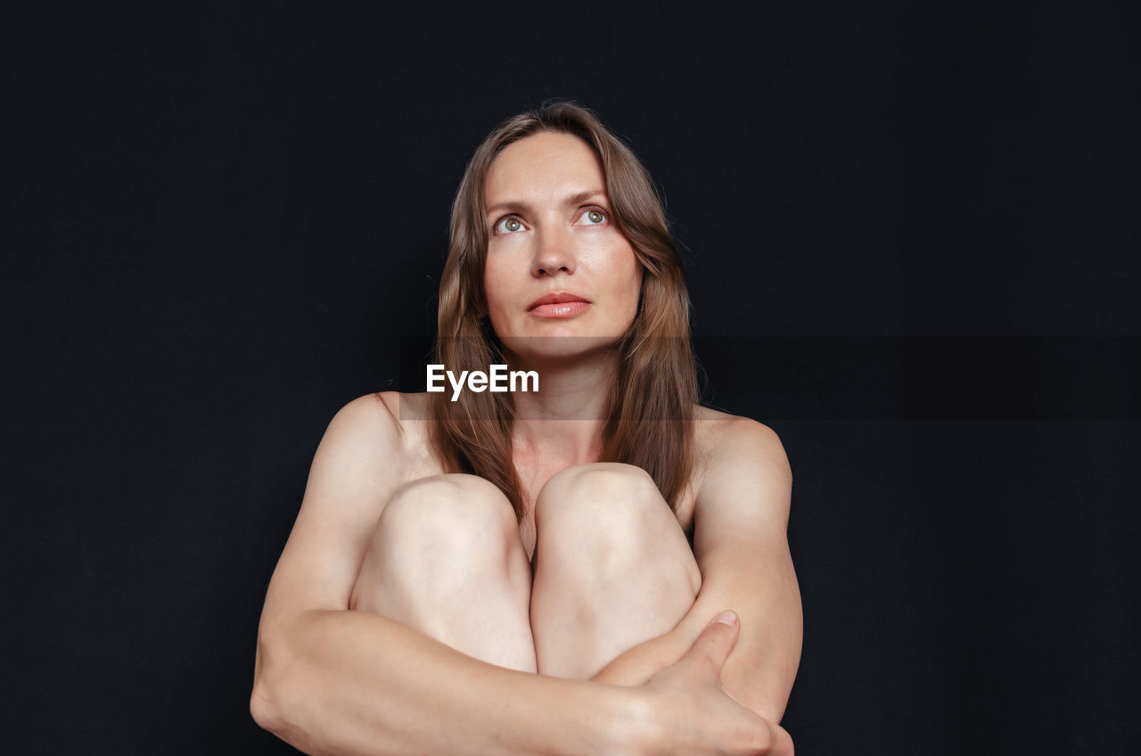 Portrait of woman on black background, sitting, hugging her knees.