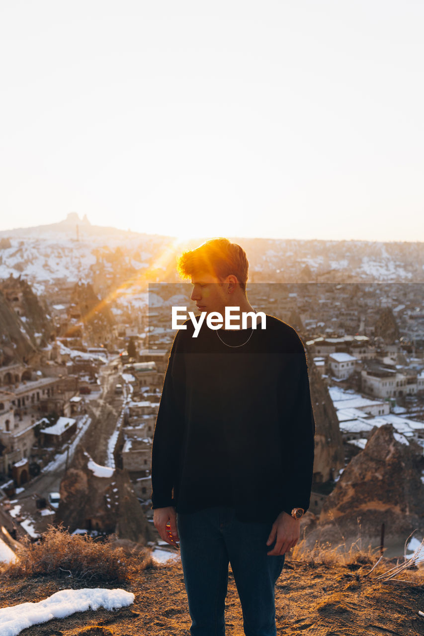 Young male in casual outfit looking away while standing against old uchisar settlement covered with snow and sunset sky in winter evening in cappadocia, turkey
