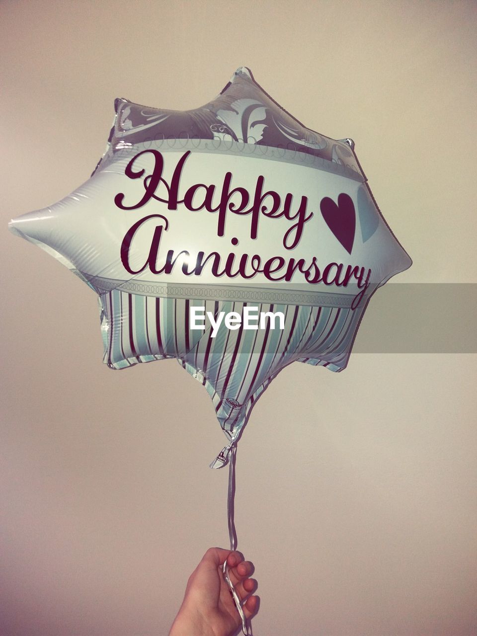 Cropped hand holding helium balloon with happy anniversary text against wall