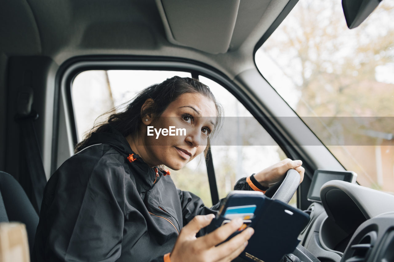 Woman holding mobile phone looking away while driving in delivery van