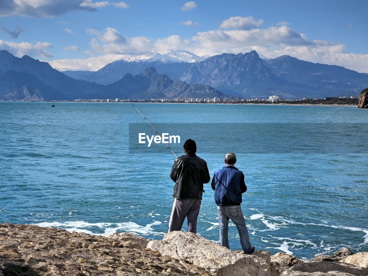 Men fishing while standing on rocky shore against mountains