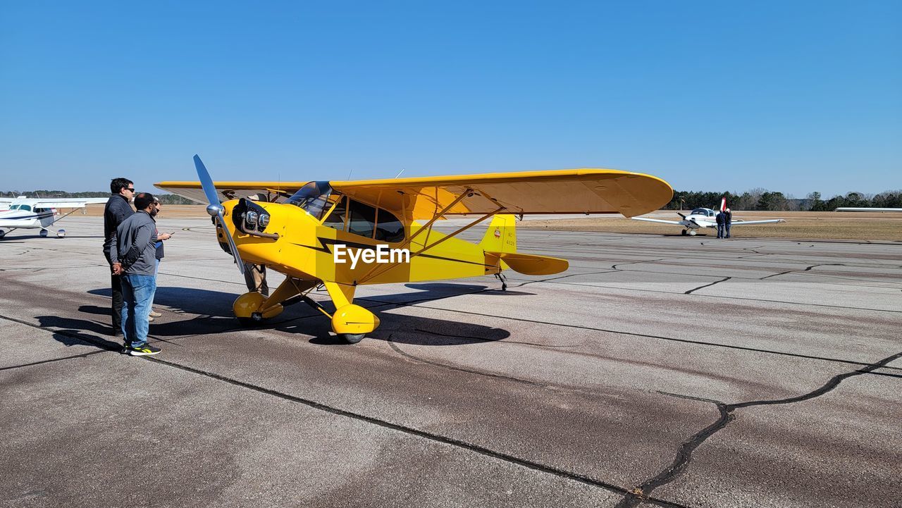 airplane, aircraft, transportation, vehicle, sky, aviation, adult, mode of transportation, clear sky, light aircraft, yellow, men, nature, sunny, sunlight, day, copy space, full length, travel, air vehicle, occupation, airport, blue, outdoors, one person, shadow, wing, working, architecture
