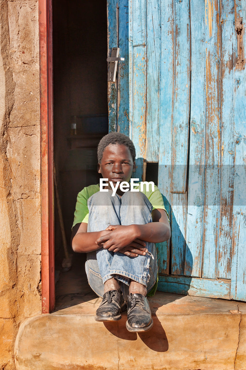 Portrait of young man hugging knees while sitting against wooden door during sunny day