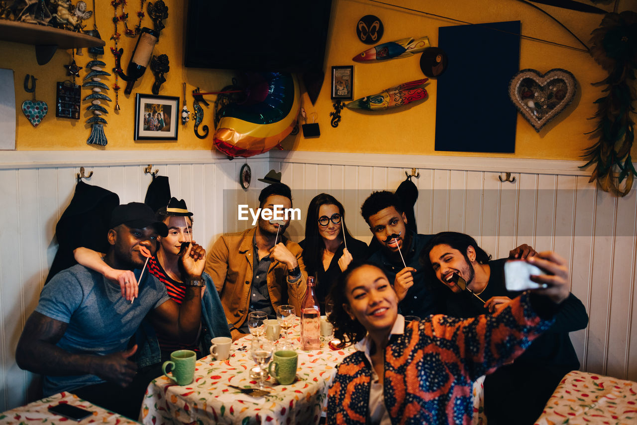 Young woman taking selfie with cheerful multi-ethnic friends holding props while sitting at restaurant during dinner par