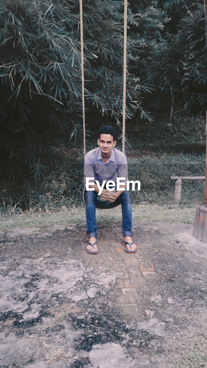 Full length portrait of smiling young man sitting on rope swing