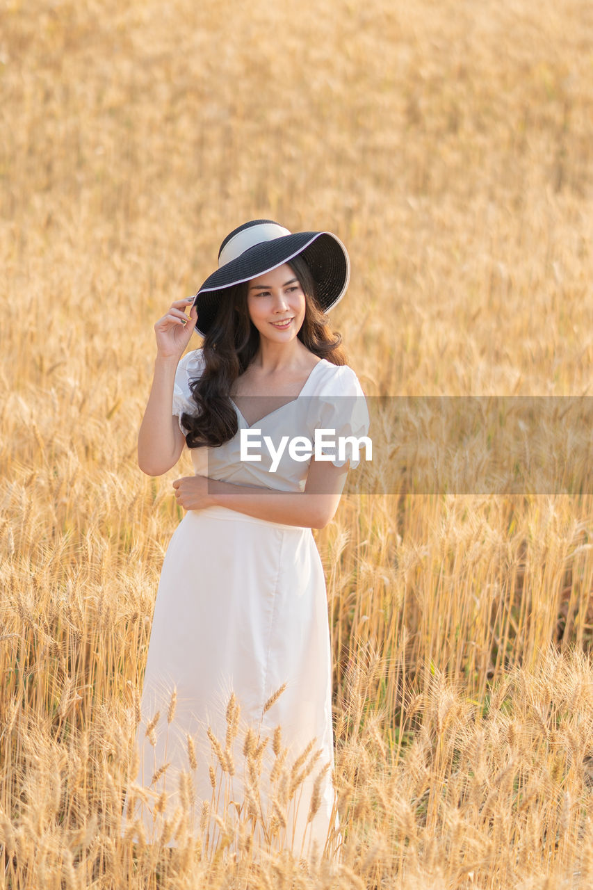 Happy young beautiful asian woman in white dress and straw hat standing in middle of barley field